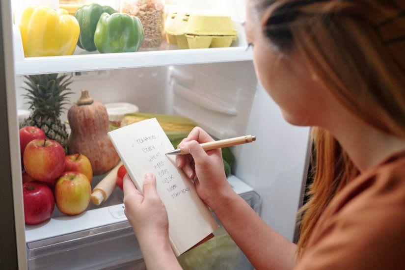 Die 48h-Regel hilft bei Impulskäufen: Lege das Produkt erst mal auf eine Wunschliste – wenn du es nach 2 Tagen immer noch brauchst, kannst du bewusst entscheiden. Frau, die am Kühlschrack eine Einkaufsliste schreibt.
